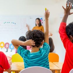 Male Students Sitting At Desk Facing The White Board With Their Hands Raised